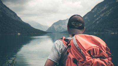 man with red hiking backpack facing body of water and mountains at daytime